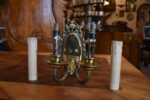 Antyczna, dwuramienna, mosiężna lampa ścienna w stylu secesyjno-klasycyzującym z owalnym szyldem i żarówkami w kształcie świec.