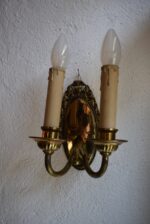 Antyczna, dwuramienna, mosiężna lampa ścienna w stylu secesyjno-klasycyzującym z owalnym szyldem i żarówkami w kształcie świec.