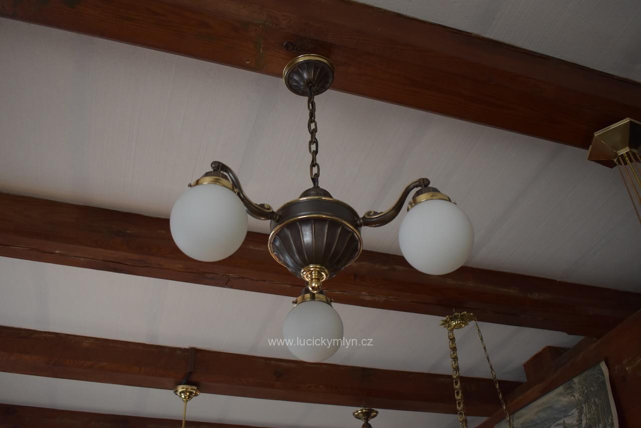 Středně velký starožitný měšťanský tří-ramenný lustr pasířsky vyrobený z tlačené a na tmavý bronz patinované, jen místy zlatě proleštěné mosazi - ztvárněno v historizujícím stylu po roce 1920 Starožitný prvorepublikový tří-ramenný lustr z patinované mosazi s mléčnými skleněnými koulemi.
