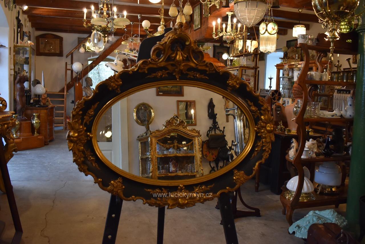 Větší starožitné oválné nástěnné zrcadlo plasticky ztvárněné v zámeckém romantickém stylu s povrchovou úpravou širokého rámu v černo‑zlatém barokizujícím až rokokově pojatém historizujícím provedení Starožitné oválné nástěnné zrcadlo v černo-zlatém rokokově historizujícím rámu