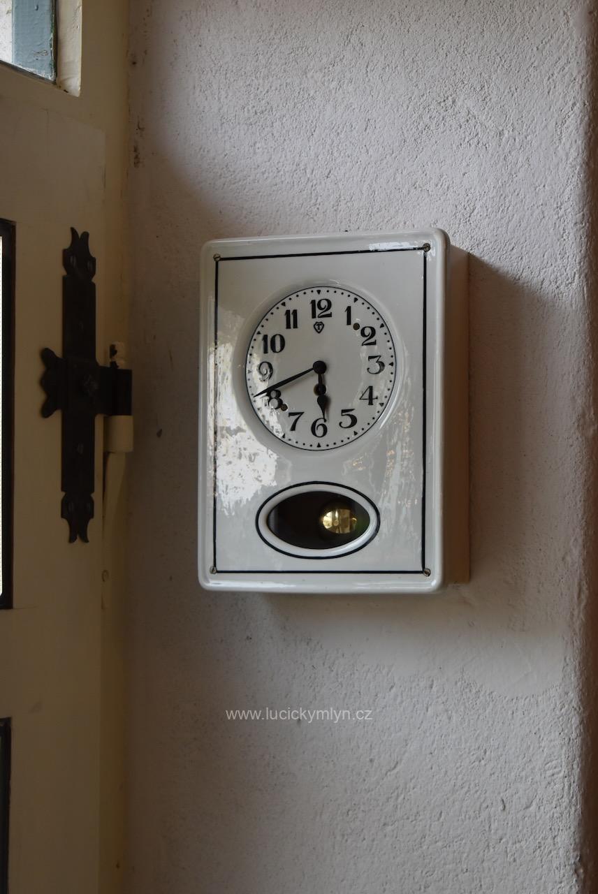 Nenápadné a decentní nástěnné kuchyňské hodiny s mechanickým pérovým strojkem usazeným ve schránce za obdélníkovým čílkem z bílého porcelánu jež má lehce zapuštěný kulatý ciferník a hladce vykrojené oválné okénko za níž se pohybuje krátké kyvadlo Starožitné prvorepublikové nástěnné kuchyňské hodiny s bílým porcelánovým čílkem a kyvadlem.