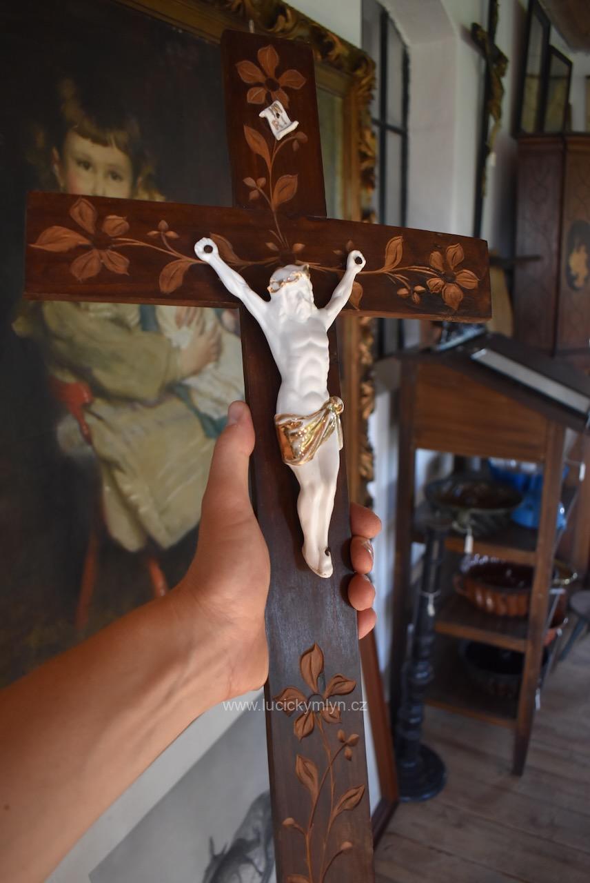 Kristus na závěsném kříži vyrobený po roce 1910 z bílého a částečně zlaceného biskvitu (neglazovaný porcelán) jež se používal pro svoji podobnost s bílým mramorem Starožitný závěsný kříž s biskvitovým Kristem na tmavě mořeném dřevě s florální řezbou.