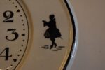 Starožitné kuchyňské hodiny s oválným porcelánovým ciferníkem a rokokovými stínovými postavičkami.