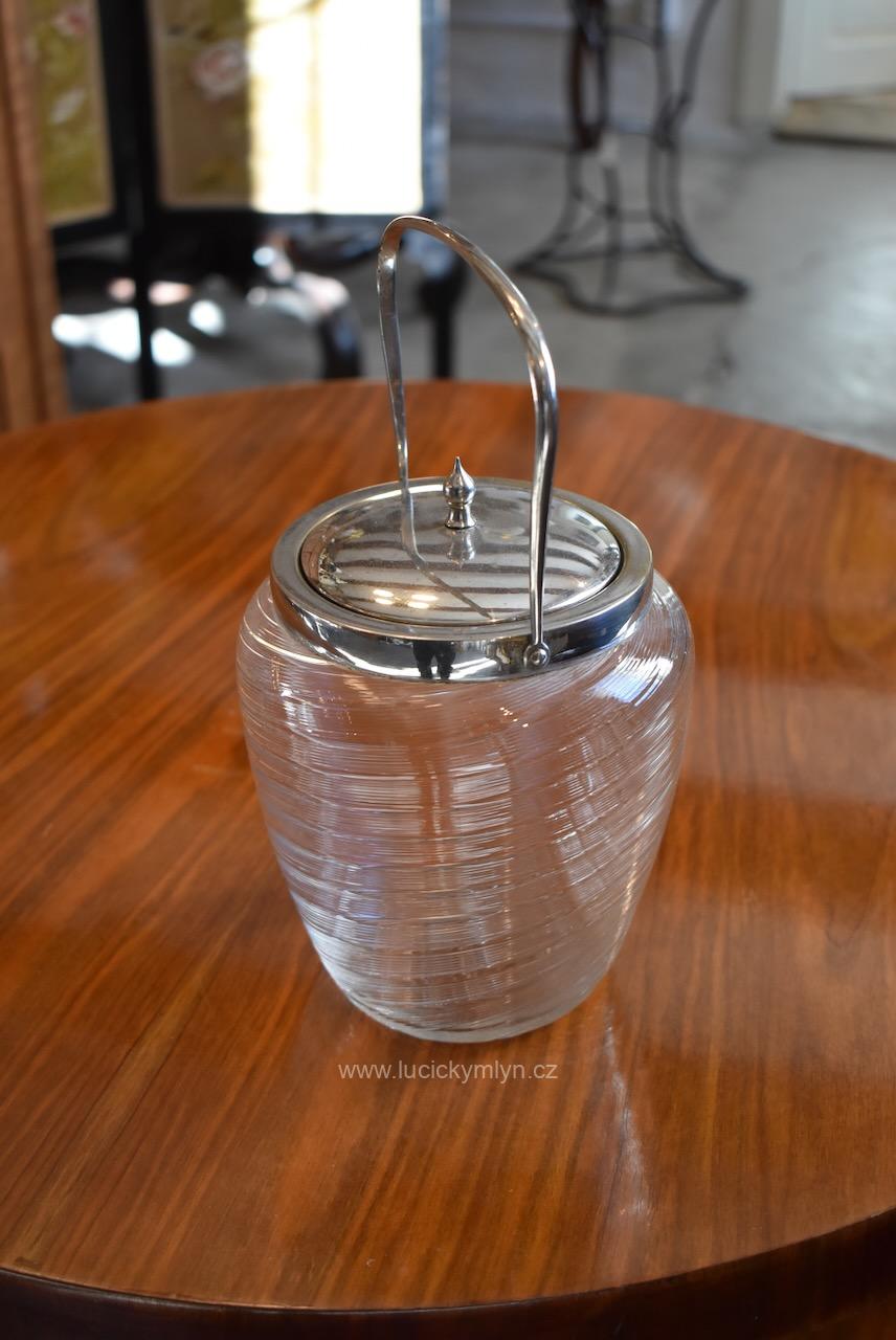 Vyšší starožitná měšťanská elegantní dóza na cukr navržená ve stylu ART-DECO z čirého foukaného skla s pláštěm spirálovitě oživeným jemnou pásovou profilací osazeno leštěnou kovovou montáží s víčkem Starožitná dóza na cukr ART-DECO z foukaného skla se stříbrně leštěnou kovovou montáží.
