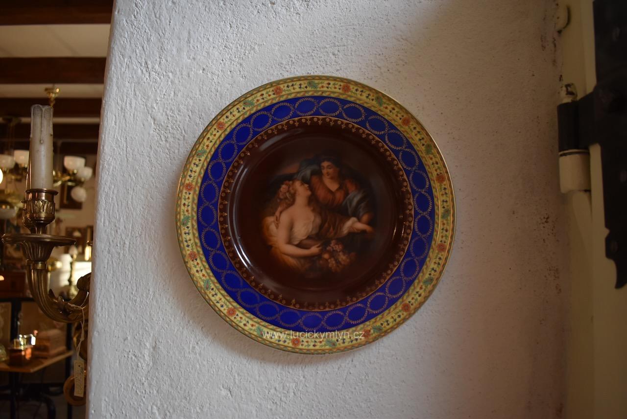 Bardzo dekoracyjna, zabytkowa wisząca paterka, malowana i ozdobiona ornamentami w stylu Royal Vienna, pochodząca z pierwszej trzeciej XX wieku. Antyczny porcelanowy talerz „Royal Vienna” z figuralnym motywem przedstawiającym dwie kobiety.